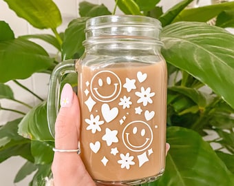 Puede incluir: Una taza de vidrio de estilo tarro de albañil con una bebida marrón en su interior. La taza tiene un diseño blanco de caras sonrientes, corazones, estrellas y diamantes.