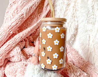 Puede incluir: Un vaso de vidrio con tapa de madera y una pajita. El vaso está decorado con un patrón de margaritas blancas sobre un fondo marrón.