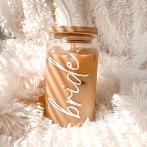 May include: A clear glass bottle with a bamboo lid and a white straw. The bottle is filled with a light brown liquid and has the word "bride" written on it in white.