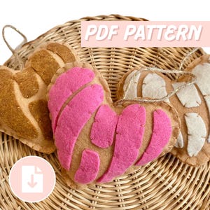 Könnte beinhalten: Drei herzförmige Filzornamente mit Concha-Stil-Designs in Braun-, Rosa- und Cremetönen, angeordnet auf einem Weidenkorb. Die Ornamente haben eine Schlaufe zum Aufhängen. Text "PDF PATTERN" ist sichtbar.