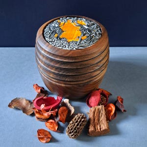 May include: A wooden box with a round lid featuring a silver embossed design of a forest scene with a yellow sun. The box is surrounded by dried flower petals and other natural elements.