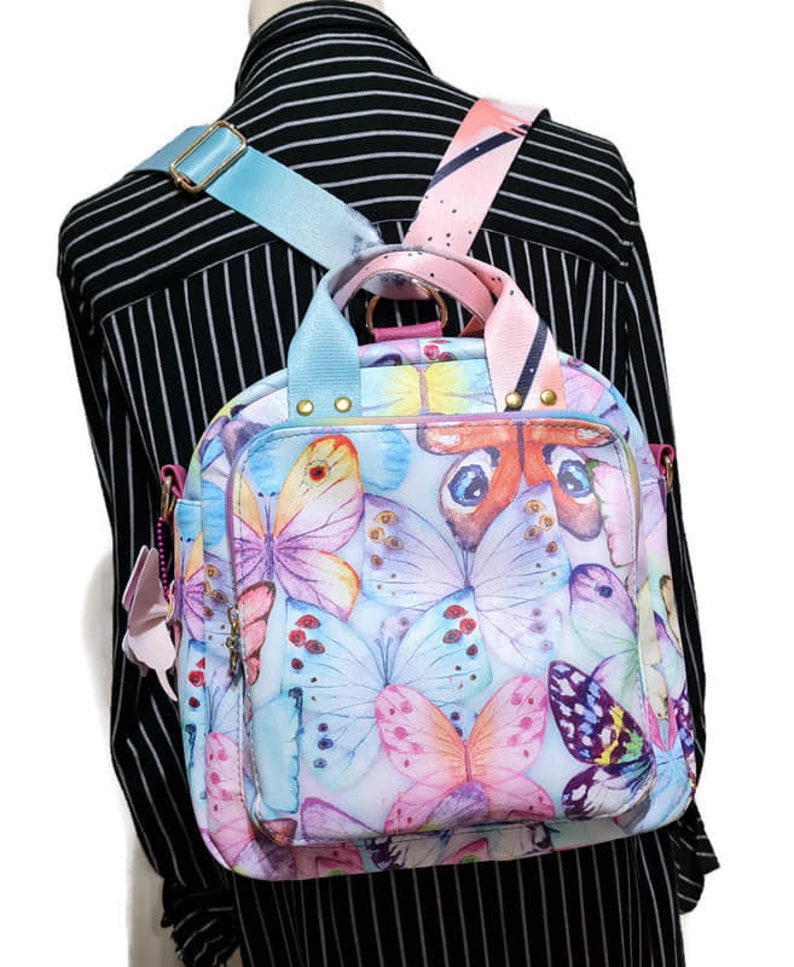May include: A light blue and pink backpack with a butterfly print. The backpack has adjustable straps and a top handle.