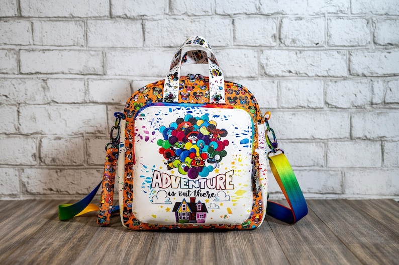 May include: White backpack with a colorful print of Disney characters and balloons. The backpack has a top handle and a long adjustable strap. The text "ADVENTURE is out there" is printed on the front of the bag.