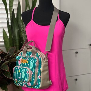 May include: A green and pink floral print backpack with a brown adjustable strap. The backpack has a zippered pocket on the front and a top handle.