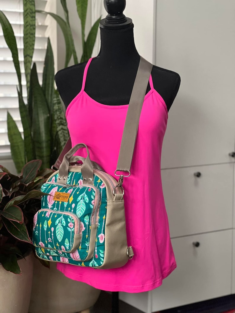 May include: A green and pink floral print backpack with a brown adjustable strap. The backpack has a zippered pocket on the front and a top handle.