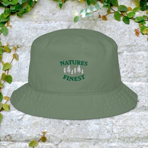 Może przedstawiać: Zielony kapelusz typu bucket hat z haftem "NATURES FINEST" z przodu. Tekst jest zielony z białym obramowaniem. Nad tekstem znajduje się pięć małych białych drzewek.