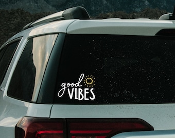 May include: White car with a black rear window and a white decal that says "good vibes" with a yellow sun graphic. The car is parked in front of a mountain range.