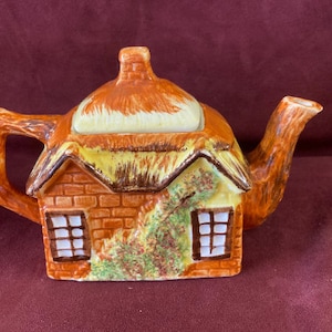 May include: A ceramic teapot shaped like a cottage with a thatched roof. The teapot features a brick facade with two windows and a climbing plant. The handle and spout are brown, and the roof is orange and yellow.
