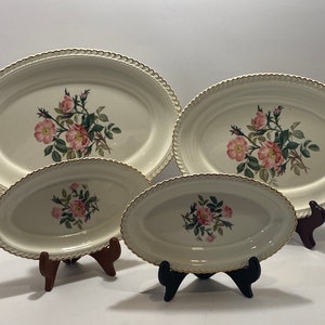 May include: Set of four oval ceramic serving dishes with a white background and a floral design featuring pink roses and green leaves. The dishes have a gold trim around the edge.