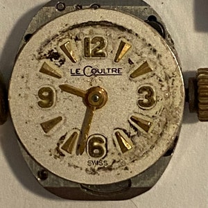 以下が含まれることがあります： 白い文字盤と金色針のヴィンテージの銀色の腕時計。時計の文字盤には黒いローマ数字の時間の目盛りと、「LE COULTRE」と「SWISS」の文字が印刷されています。
