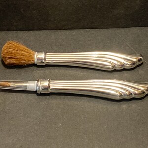 May include: Two silver-toned makeup brushes with a decorative, fluted handle. The brush on the top has a brown bristle brush head. The brush on the bottom has a clear plastic tube with a silver-toned tip.