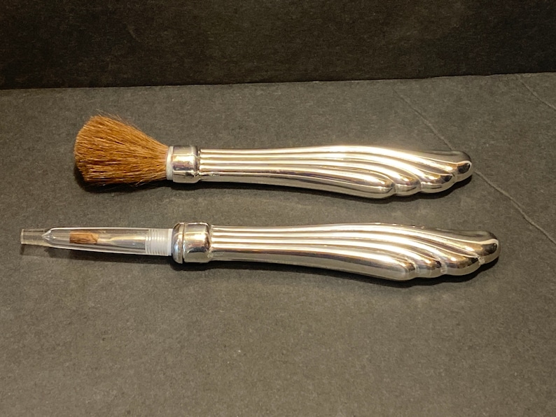 May include: Two silver-toned makeup brushes with a decorative, fluted handle. The brush on the top has a brown bristle brush head. The brush on the bottom has a clear plastic tube with a silver-toned tip.
