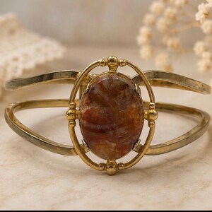 May include: A gold-toned metal cuff bracelet featuring a central oval-shaped stone with a marbled brown and amber pattern. The stone is encased in a decorative oval frame with small spherical accents, connected to a split-band cuff design.