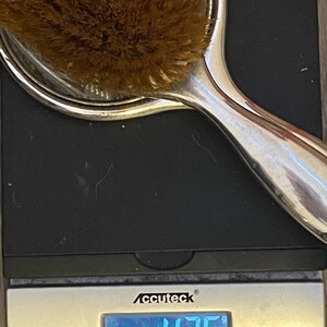 Puede incluir: Un espejo de mano plateado con un cepillo marr&oacute;n en la parte delantera. El espejo est&aacute; en una b&aacute;scula digital que marca 426 gramos.