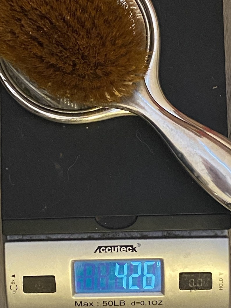 Puede incluir: Un espejo de mano plateado con un cepillo marr&oacute;n en la parte delantera. El espejo est&aacute; en una b&aacute;scula digital que marca 426 gramos.