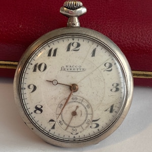 May include: A silver pocket watch with Roman numeral markings on the face. The watch face also includes the text "RACCO LEVRETTE" and a smaller circular dial with black markings.