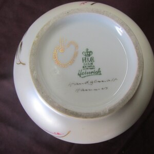May include: The bottom of a white porcelain vase with a gold and green mark that reads "H&C Selb Bavaria Germany  Heubrich" and a handwritten inscription that reads "Handgemalt  Hahnennest".