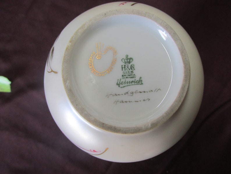 May include: The bottom of a white porcelain vase with a gold and green mark that reads "H&C Selb Bavaria Germany  Heubrich" and a handwritten inscription that reads "Handgemalt  Hahnennest".