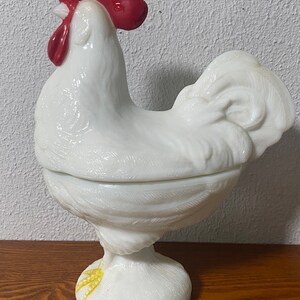 May include: White milk glass rooster shaped candy dish with a red comb and yellow feet. The dish has a lid and is a vintage item.