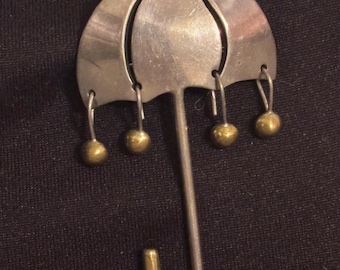 May include: A silver-toned metal umbrella-shaped brooch with a triangular top and curved ribs. Four small gold-colored spheres dangle from the umbrella's edge. The handle has a hook at the end, also gold-colored.