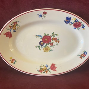May include: Oval serving platter with a white background and a red rim. The platter is decorated with colorful floral arrangements in red, blue, yellow, and green. A classic piece for serving food.