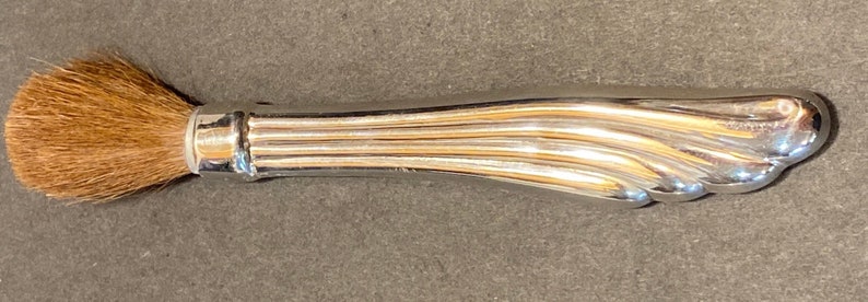May include: A silver makeup brush with a brown bristle brush head. The handle is fluted and has a rounded end.