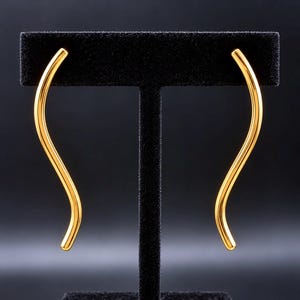 May include: A pair of gold-toned earrings, each featuring a smooth, elongated, and slender S-shaped curve. The metal surface is polished and reflective, showcasing a continuous, flowing design that tapers slightly at both ends.