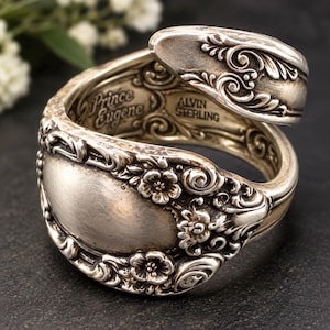 May include: A silver spoon ring featuring an intricate floral pattern and scrollwork. The interior of the band is clearly engraved with the text Prince Eugene and ALVIN STERLING, showcasing the detailed craftsmanship of this metal accessory.