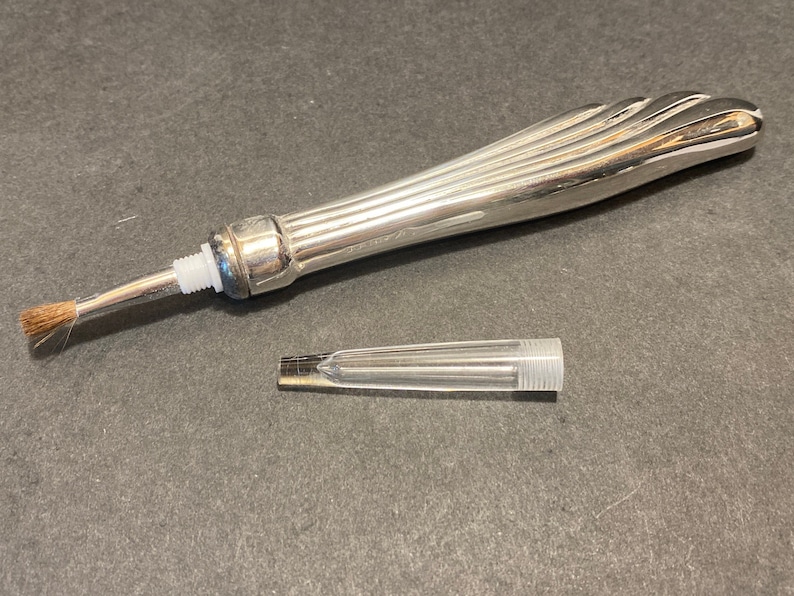 May include: A silver-toned brush with a long, fluted handle and a small, clear plastic tube. The brush has a small, brown bristle head.