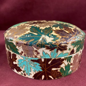 May include: A round, vintage trinket box with a white background and a pattern of green, brown, and gold leaves. The box is made of paper mache and has a slightly worn appearance.