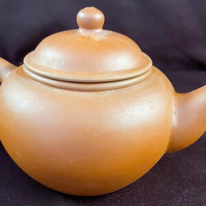 May include: A small, brown ceramic teapot with a lid. The teapot has a round body and a short spout.