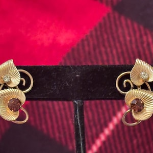 May include: Gold-toned leaf-shaped earrings with a vintage aesthetic. Each earring features two textured leaves, one with a small clear stone and the other with a dark brown stone. The earrings are displayed on a black stand against a red and black plaid background.