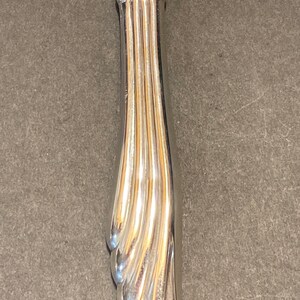 May include: Silver-toned makeup brush handle with a decorative, fluted design. The handle is curved and has a rounded tip.
