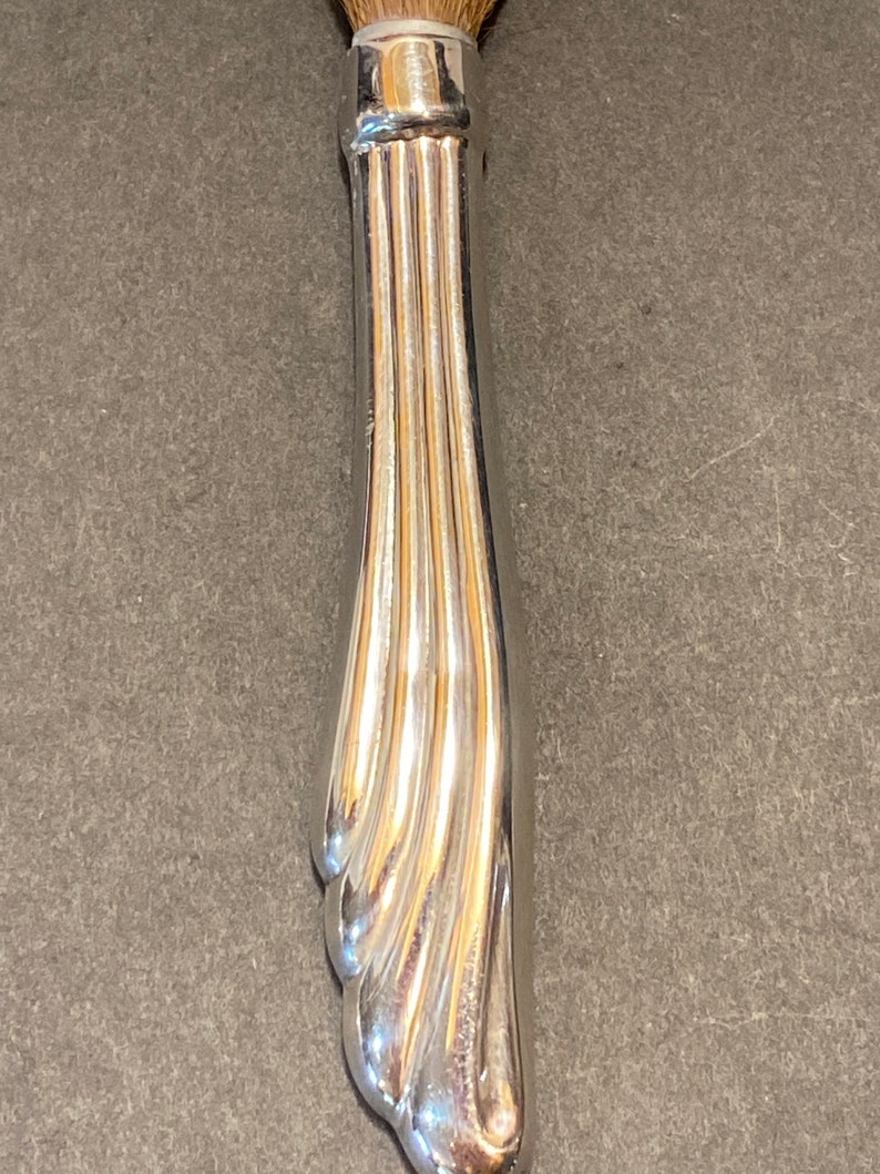 May include: Silver-toned makeup brush handle with a decorative, fluted design. The handle is curved and has a rounded tip.