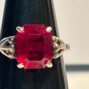 May include: A silver ring featuring a large, rectangular, deep red gemstone. The ring has a detailed, ornate design on the band with small, round accents on either side of the gemstone. The gemstone is held in place by four small prongs.