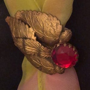 May include: A gold-toned metal napkin ring with a leaf design and a large, faceted red jewel. The napkin ring is wrapped around a sheer fabric in shades of yellow and pink.