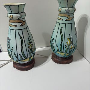 May include: A pair of ceramic table lamps with a blue and green undersea theme. The lamps feature a variety of sea creatures, including fish, seaweed, and shells. The lamps are on wooden bases.