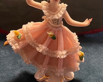 May include: A porcelain figurine of a woman in a pink and white dress with lace trim. She is holding her arms out and has a delicate, feminine look.