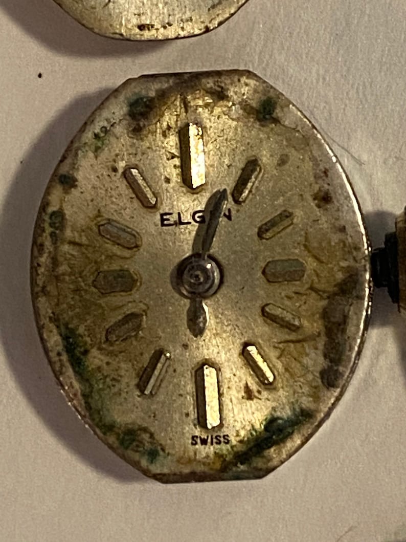 以下が含まれることがあります： 銀色の時計の文字盤のクローズアップ。黒い針とローマ数字の時間の目盛りがあります。文字盤には「ELGIN」と「SWISS」と書かれています。