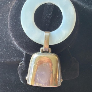 May include: A vintage pendant featuring a silver-toned bell charm suspended from a pale blue, circular ring. The bell has a reflective surface and a rounded shape. The ring is attached to the bell with a small metal clasp. The pendant is displayed on a black surface.