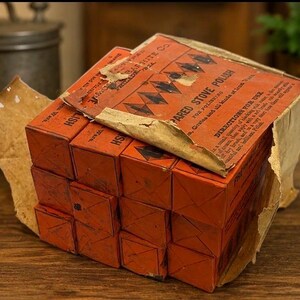 May include: A stack of rectangular orange boxes labeled Prepared Stove Polish by the Graphite Co. The packaging features black diamond graphics and printed text with directions for use. The paper wrapping is torn and worn, revealing the stacked boxes.