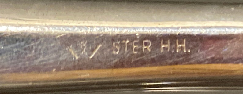 May include: Close-up of a silver-toned object with the inscription "STER H.H."