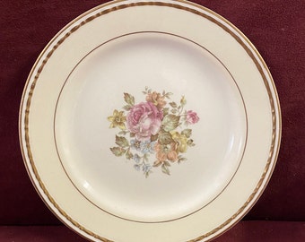 May include: A white ceramic dinner plate with a gold rim and a floral design in the center. The flowers are pink, yellow, and blue.
