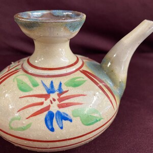 May include: A white ceramic sake bottle with a brown, green, red, and blue floral design. The bottle has a spout and a wide opening.