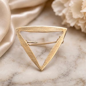 May include: A gold-toned metal brooch in the shape of an open triangle. The surface of the brooch features a brushed, matte texture. A thin metal pin mechanism is visible across the center of the triangular frame, extending from the left side.