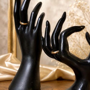 May include: Two smooth, black, upright hand-shaped jewelry displays. The taller, vertical hand model wears a simple gold band on its ring finger, while the shorter, horizontal hand model wears a matching gold band on its ring finger.