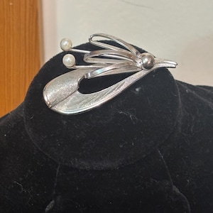 May include: A silver-toned brooch with a leaf-like design and two small pearl accents. The brooch has a textured finish and a curved, flowing shape. The brooch is set against a black velvet background.