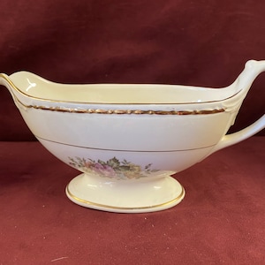 May include: A white ceramic gravy boat with a gold trim and floral design. The boat has a handle and a pedestal base.
