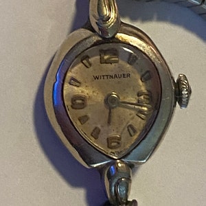 May include: A vintage Wittnauer wristwatch with a silver-toned case and a white face. The watch has a delicate, oval-shaped design and a metal band.