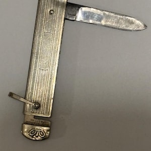 May include: A silver pocket knife with a partially open blade. The knife has a ribbed design on the handle and a decorative floral pattern at the base. The blade is silver and shows signs of wear.